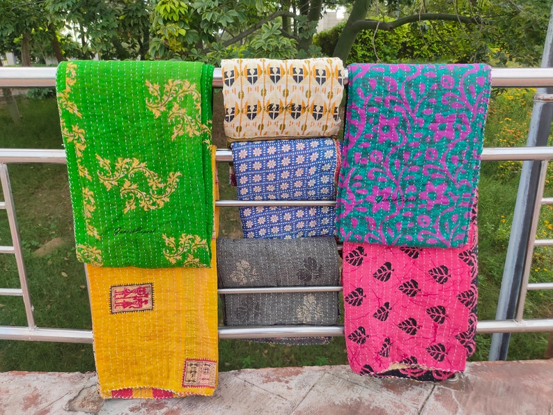 May include: Five kantha quilts hanging on a metal rack. The quilts are made of cotton and have different patterns and colours, including green, yellow, blue, pink, and black.