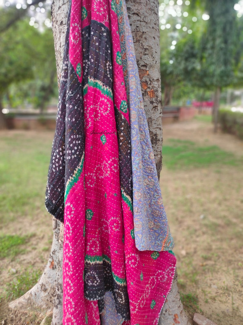 May include: A pink, black and blue kantha quilt with a floral pattern. The quilt is draped over a tree trunk.