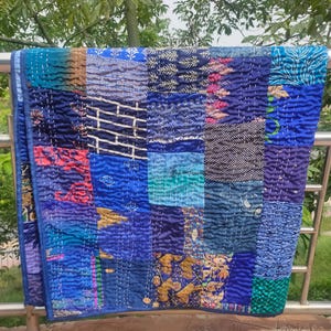 May include: A patchwork quilt with a variety of blue fabrics, including teal, navy, and royal blue. The quilt is made of squares and rectangles of different patterns and textures, and is hand-stitched with white thread. The quilt is draped over a metal railing.