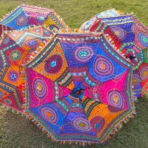 Puede incluir: Un grupo de coloridos paraguas decorativos abiertos con intrincados patrones geométricos. Los paraguas presentan tonos vibrantes de naranja, rosa, azul y morado, con diseños circulares detallados y bordes con borlas. Están dispuestos sobre una superficie cubierta de hierba.