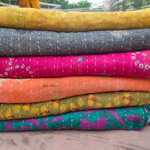 May include: A stack of six kantha quilts in various colours and patterns, including yellow, grey, pink, orange, green, and purple. The quilts are made from recycled saris and are hand-stitched.