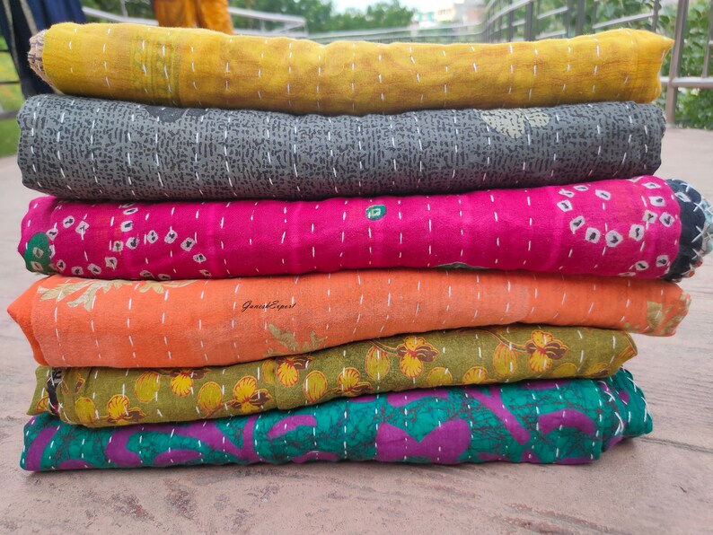 May include: A stack of six kantha quilts in various colours and patterns, including yellow, grey, pink, orange, green, and purple. The quilts are made from recycled saris and are hand-stitched.