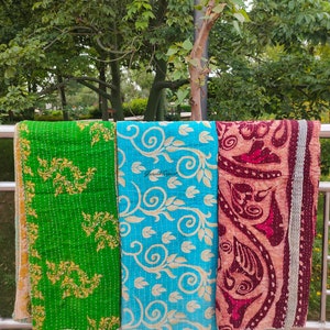 May include: Three kantha quilts hanging on a railing. The quilts are made of cotton and have a variety of patterns. The quilt on the left is green with gold floral designs. The quilt in the middle is blue with white floral designs. The quilt on the right is maroon with white floral designs.