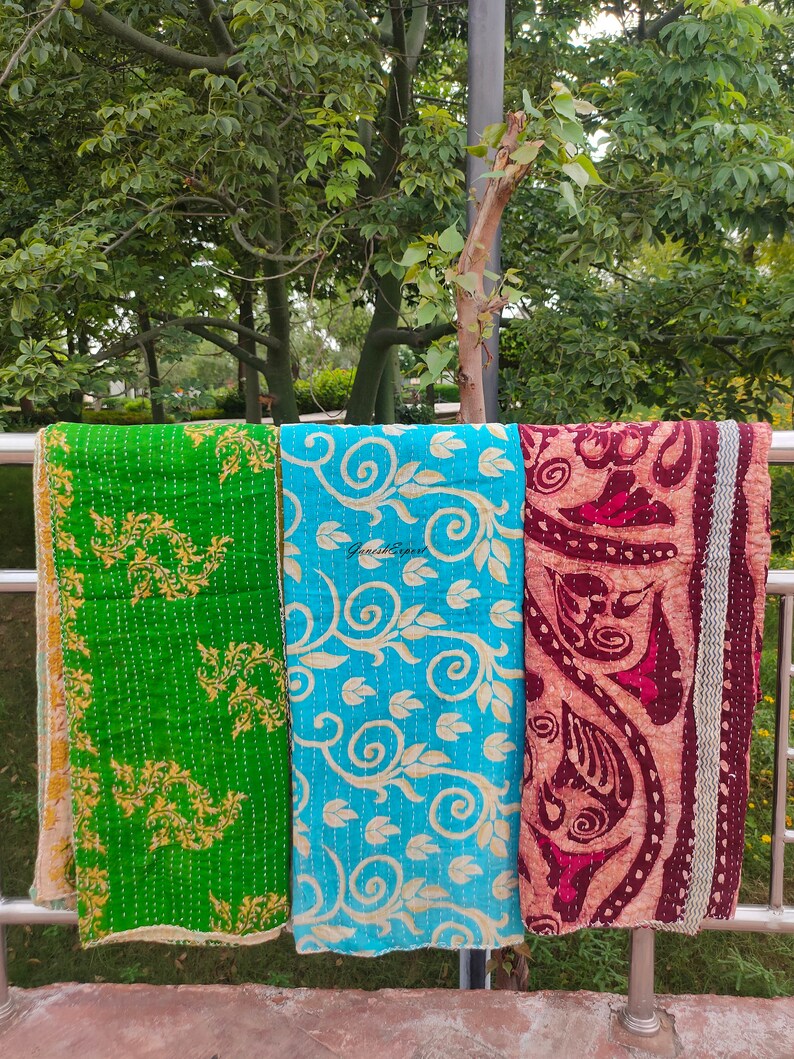 May include: Three kantha quilts hanging on a railing. The quilts are made of cotton and have a variety of patterns. The quilt on the left is green with gold floral designs. The quilt in the middle is blue with white floral designs. The quilt on the right is maroon with white floral designs.