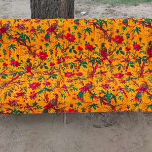 May include: A yellow kantha quilt with a floral and bird pattern in red, green, and yellow. The quilt is folded and placed on a green metal bench.