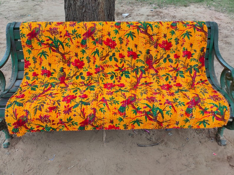 May include: A yellow kantha quilt with a floral and bird pattern in red, green, and yellow. The quilt is folded and placed on a green metal bench.