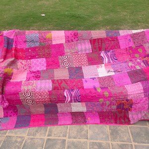 May include: A rectangular patchwork quilt featuring a variety of pink, magenta, and purple fabric squares. The quilt displays diverse patterns including geometric shapes, swirls, and floral motifs, all joined with visible running stitch embroidery.