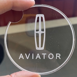 May include: Clear acrylic car coaster with the word "AVIATOR" and a stylized emblem etched into the surface. The coaster is round with a small notch for easy removal from a car cup holder.
