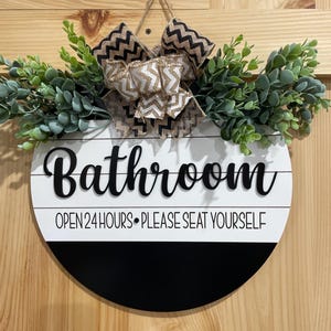 May include: Round wooden bathroom sign with a black and white design. The sign has the word "Bathroom" in a cursive font, with the text "OPEN 24 HOURS • PLEASE SEAT YOURSELF" below. It is decorated with greenery and a bow.