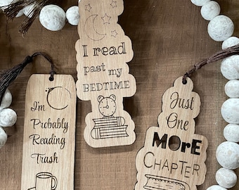 May include: Three wooden bookmarks with different sayings. The first bookmark says "I'm Probably Reading Trash" and has a coffee cup and book illustration. The second bookmark says "I read past my bedtime" and has a clock and stack of books illustration. The third bookmark says "Just One More Chapter" and has a stack of books illustration.