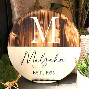 May include: A round wooden sign with a white background and a burnt wood top. The sign features a large white letter "M" and the text "Malzahn EST. 1995" in black.
