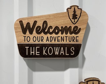 May include: Wooden sign with the words "Welcome to our Adventure" and "The Kowals" engraved in dark brown. A shield-shaped emblem with a tree is on the top right. The sign is made of light-colored wood.