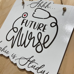 May include: A white square wooden sign with rounded corners and a jute twine hanger. The sign features black text that reads "Shhh... FUTURE Nurse" and "Sophie is Studying" with a black stethoscope graphic and a small red heart icon.