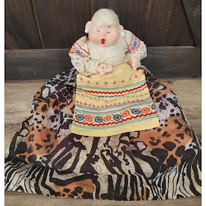 May include: A vintage doll with a painted face, wearing a cream-colored blouse with red and blue patterned sleeves. The doll has a yellow apron with red, blue, and green embroidery. The skirt is a mix of animal prints in brown, black, and white.