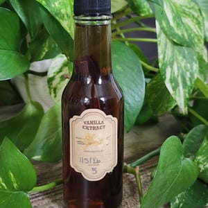 Könnte beinhalten: Eine klare Glasflasche gefüllt mit dunkelbraunem Vanilleextrakt, verschlossen mit einem schwarzen Schraubdeckel. Das Etikett trägt die Aufschrift VANILLA EXTRACT, HANDCRAFTED BY, DATE 11/5/26 und BATCH NO 5 sowie eine Abbildung einer Vanilleblüte.