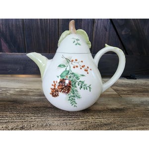 Può includere: Una teiera in ceramica a forma di pera con base color crema, caratterizzata da un disegno dipinto di pigne marroni, aghi di pino verdi e bacche arancioni. Il coperchio ha la forma di un picciolo di pera con foglie verde chiaro.
