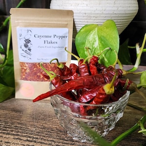 May include: A clear glass bowl filled with red chili peppers sits in front of a package of cayenne pepper flakes. The package has the text "Cayenne Pepper Flakes - Farm Fresh Ingredients". A green leafy plant and a white vase are in the background.