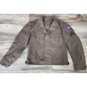 May include: A vintage olive-brown military jacket with four front pockets and a button closure. The jacket features a patch on the upper left sleeve with a red, white, and blue emblem and a green chevron. The jacket is displayed on a wooden surface.