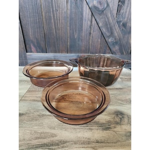 Op de afbeelding: Drie amberkleurige glazen kookgerei. De set bevat twee ronde schalen en een kom met handvatten. Het glas heeft een geribbeld ontwerp. Het kookgerei is geplaatst op een houten oppervlak.