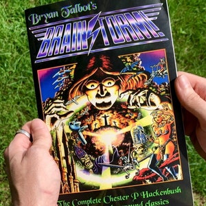 May include: A comic book cover for "Brainstorm" by Bryan Talbot, held in hands. The cover features a vibrant, surreal illustration with a central face, and the title in a stylized font. The text "The Complete Chester P Hackenbush and other underground classics" is also visible.