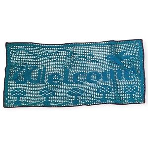 May include: A teal blue crocheted table runner with the word "Welcome" in the center. The runner has a delicate, lacy pattern.