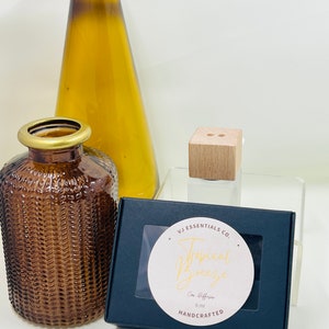 Puede incluir: Una caja negra con una ventana transparente que muestra un círculo blanco con el texto "VJ Essentials Co. Tropical Breeze Car Diffuser 6 ml HANDCRAFTED". La caja está sobre una superficie transparente junto a una botella de vidrio marrón y un cubo de madera.