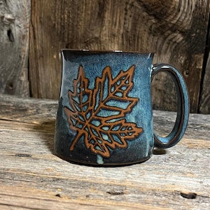 May include: A ceramic mug with a blue glaze and a brown maple leaf design. The mug has a dark brown handle and rim. The mug is sitting on a wooden surface.