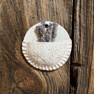 Pu&ograve; includere: Una piccola tasca da parete in ceramica bianca macchiettata con un bordo smerlato e un motivo floreale in rilievo. La tasca contiene un mazzetto di lavanda essiccata. La tasca da parete &egrave; montata su una superficie di legno rustico.