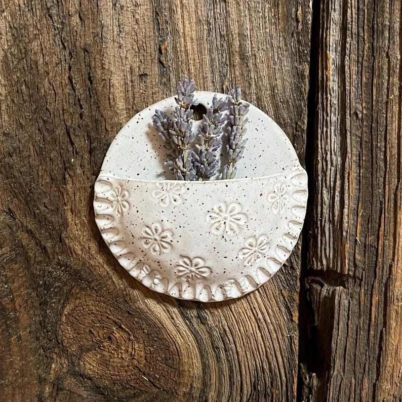 Pu&ograve; includere: Una piccola tasca da parete in ceramica bianca macchiettata con un bordo smerlato e un motivo floreale in rilievo. La tasca contiene un mazzetto di lavanda essiccata. La tasca da parete &egrave; montata su una superficie di legno rustico.
