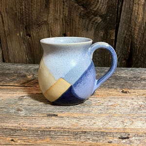 May include: A handmade ceramic mug with a rounded body and a curved handle. The mug features a geometric design with shades of blue, tan, and navy. The mug is sitting on a weathered wooden surface.