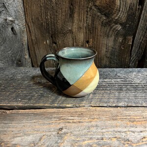 May include: A handmade ceramic mug with a black handle and a colorful geometric design. The mug features sections of black, tan, yellow, and teal. The mug is sitting on a wooden surface.