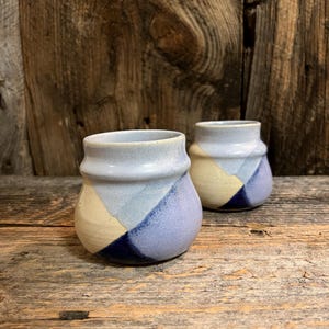 May include: Two small, handmade ceramic vases with a geometric design. The vases feature a color block pattern in shades of light blue, cream, and navy blue. The vases are displayed on a rustic wooden surface.