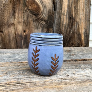 May include: A small, blue ceramic jar with a textured rim and two brown leaf-like designs. The jar sits on a weathered wooden surface, with a rustic wooden background.