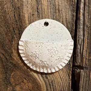 Pu&ograve; includere: Una tasca da parete in ceramica bianco sporco macchiettata, a forma di semicerchio. La met&agrave; inferiore presenta motivi floreali in rilievo e un bordo smerlato. C'&egrave; un piccolo foro nella parte superiore per appendere.