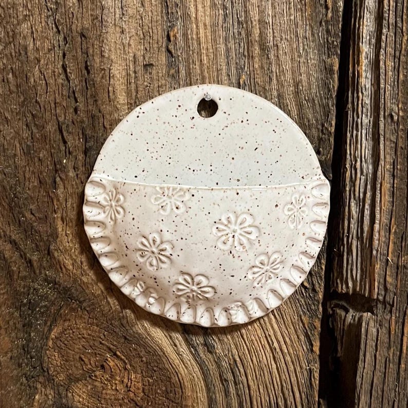 Pu&ograve; includere: Una tasca da parete in ceramica bianco sporco macchiettata, a forma di semicerchio. La met&agrave; inferiore presenta motivi floreali in rilievo e un bordo smerlato. C'&egrave; un piccolo foro nella parte superiore per appendere.