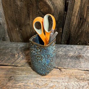 May include: A ceramic pen holder with a textured blue glaze, holding orange scissors and two gray pens. The pen holder has a unique, irregular shape and sits on a wooden surface.