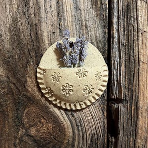 May include: A beige ceramic wall pocket with a scalloped edge and embossed floral designs. The pocket holds dried lavender sprigs. The wall hanging is mounted on a weathered wooden surface, creating a rustic aesthetic.