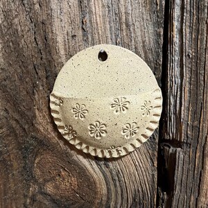 Pu&ograve; includere: Una tasca da parete in ceramica beige a forma di semicerchio. La met&agrave; inferiore presenta motivi floreali in rilievo e un bordo smerlato. Un piccolo foro nella parte superiore per appenderla. Su uno sfondo di legno rustico.