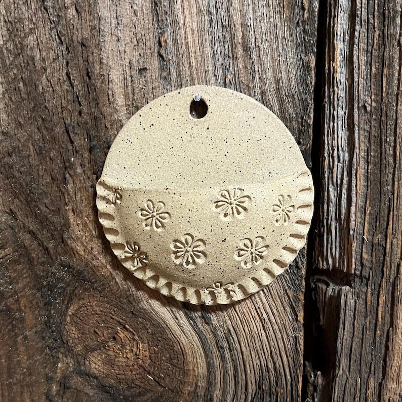 Pu&ograve; includere: Una tasca da parete in ceramica beige a forma di semicerchio. La met&agrave; inferiore presenta motivi floreali in rilievo e un bordo smerlato. Un piccolo foro nella parte superiore per appenderla. Su uno sfondo di legno rustico.