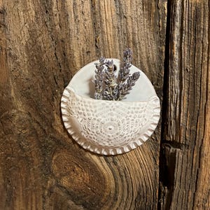 May include: A small, white ceramic wall pocket with a lace-like pattern and a ruffled edge, holding dried lavender sprigs. The pocket is mounted on a weathered, dark wood surface, creating a rustic aesthetic.