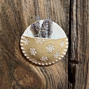 Pu&ograve; includere: Una tasca da parete in ceramica fatta a mano con un motivo floreale marrone chiaro e una sezione superiore bianca. La tasca contiene rametti di lavanda essiccati. Il pezzo &egrave; montato su uno sfondo di legno rustico, creando un'estetica naturale e terrosa.