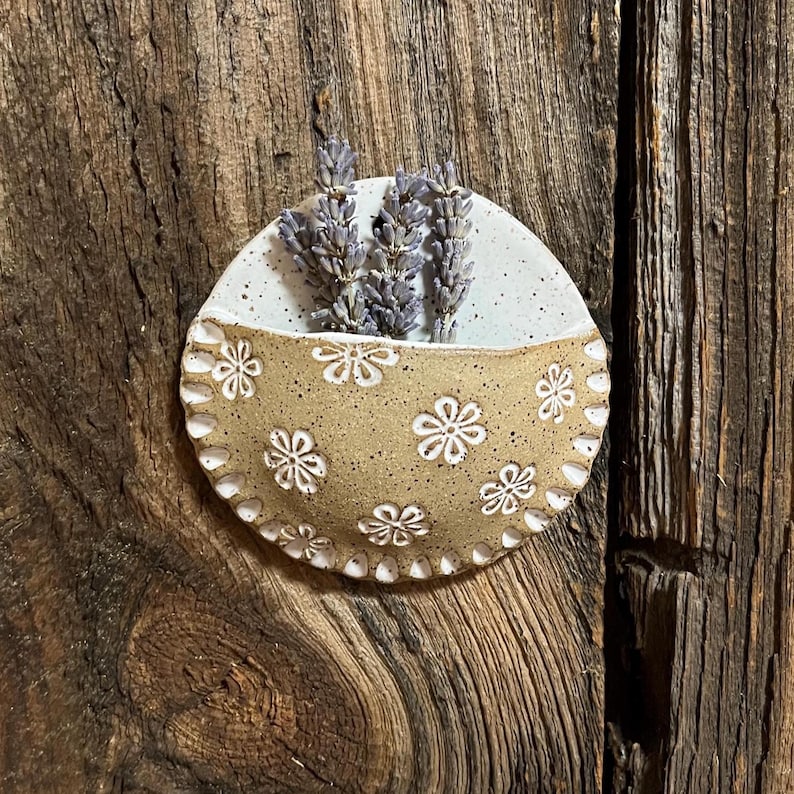 Pu&ograve; includere: Una tasca da parete in ceramica fatta a mano con un motivo floreale marrone chiaro e una sezione superiore bianca. La tasca contiene rametti di lavanda essiccati. Il pezzo &egrave; montato su uno sfondo di legno rustico, creando un'estetica naturale e terrosa.