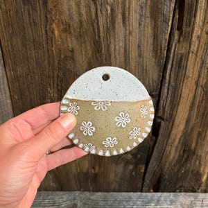 Pu&ograve; includere: Una tasca da parete in ceramica fatta a mano con un design a semicerchio. La met&agrave; superiore &egrave; bianca e la met&agrave; inferiore &egrave; beige con motivi floreali bianchi. Ha un piccolo foro per appenderla. Il pezzo &egrave; tenuto in mano.