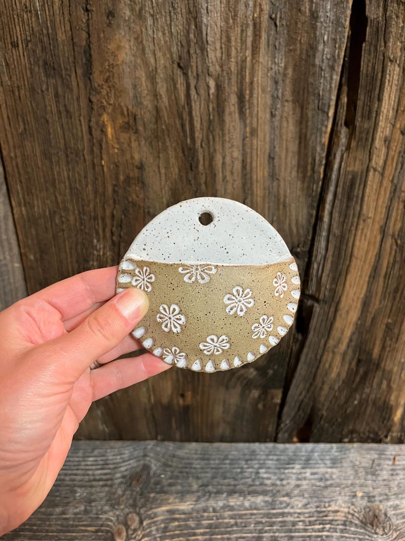 Pu&ograve; includere: Una tasca da parete in ceramica fatta a mano con un design a semicerchio. La met&agrave; superiore &egrave; bianca e la met&agrave; inferiore &egrave; beige con motivi floreali bianchi. Ha un piccolo foro per appenderla. Il pezzo &egrave; tenuto in mano.