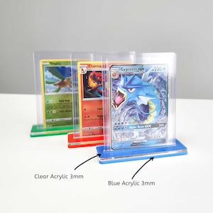 May include: Three clear acrylic card display stands with a 3mm thick base. The stands are in different colors: green, red, and blue. The stands are holding trading cards featuring Pokemon characters, including Tropius, Charmeleon, and Gyarados.