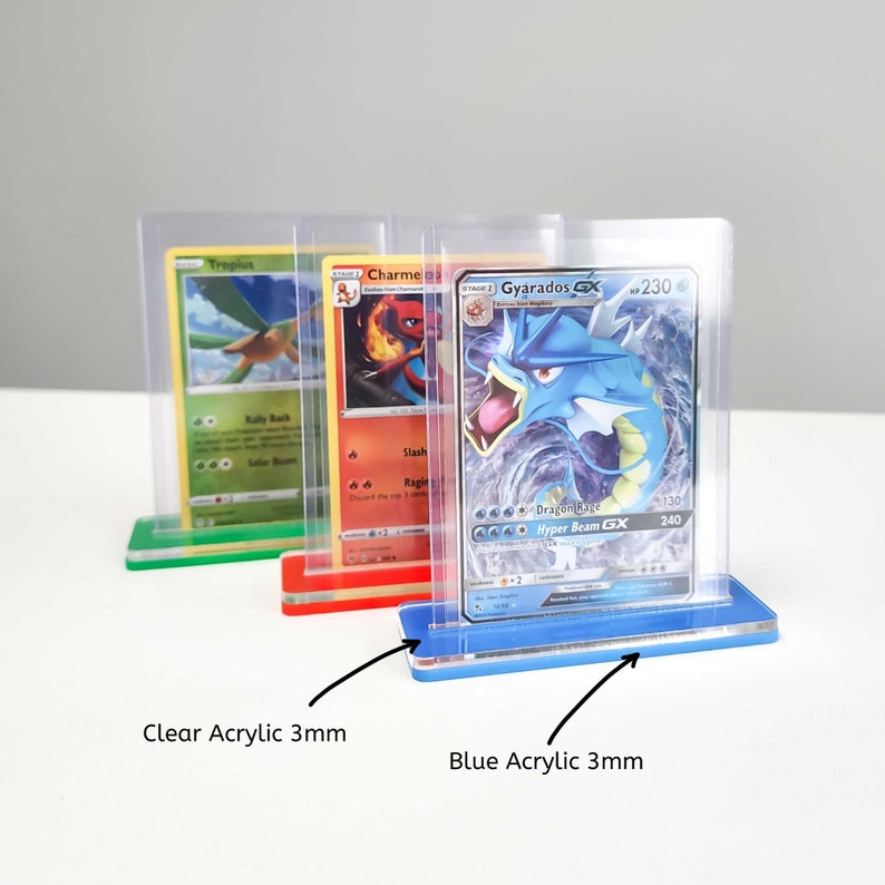 May include: Three clear acrylic card display stands with a 3mm thick base. The stands are in different colors: green, red, and blue. The stands are holding trading cards featuring Pokemon characters, including Tropius, Charmeleon, and Gyarados.