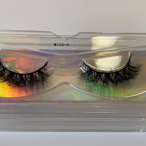 May include: A pair of black false eyelashes in a clear plastic case. The lashes are thick and voluminous, with a curved shape. The case has a holographic iridescent background and the text "M100-P".