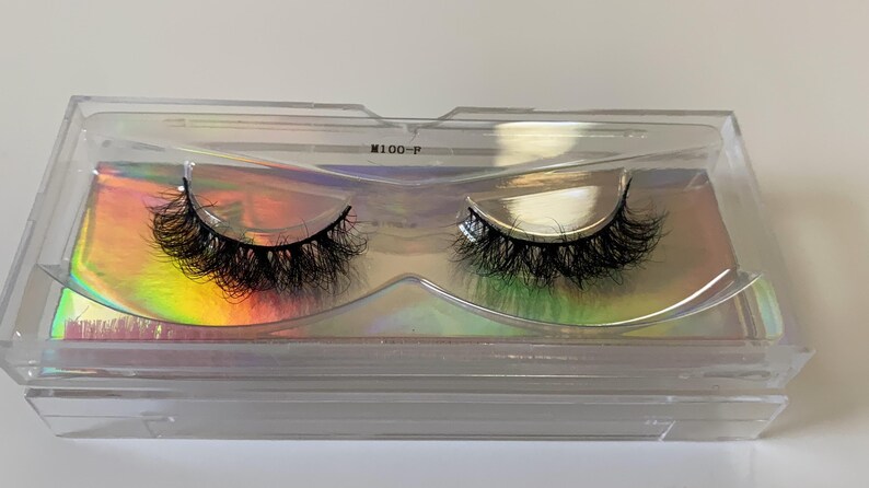 May include: A pair of black false eyelashes in a clear plastic case. The lashes are thick and voluminous, with a curved shape. The case has a holographic iridescent background and the text "M100-P".