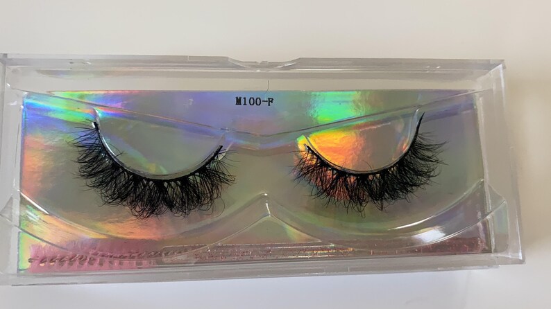 May include: A pair of black false eyelashes in a clear plastic case. The case has a holographic background with the text "M100-P". The eyelashes are long and full, designed to enhance the eyes.
