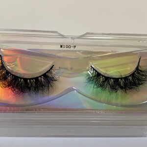 May include: A pair of black false eyelashes in a clear plastic case. The lashes are thick and fluffy, with a natural-looking curl. The case has a holographic insert that reflects a rainbow of colors. The text "M100-F" is printed on the case.